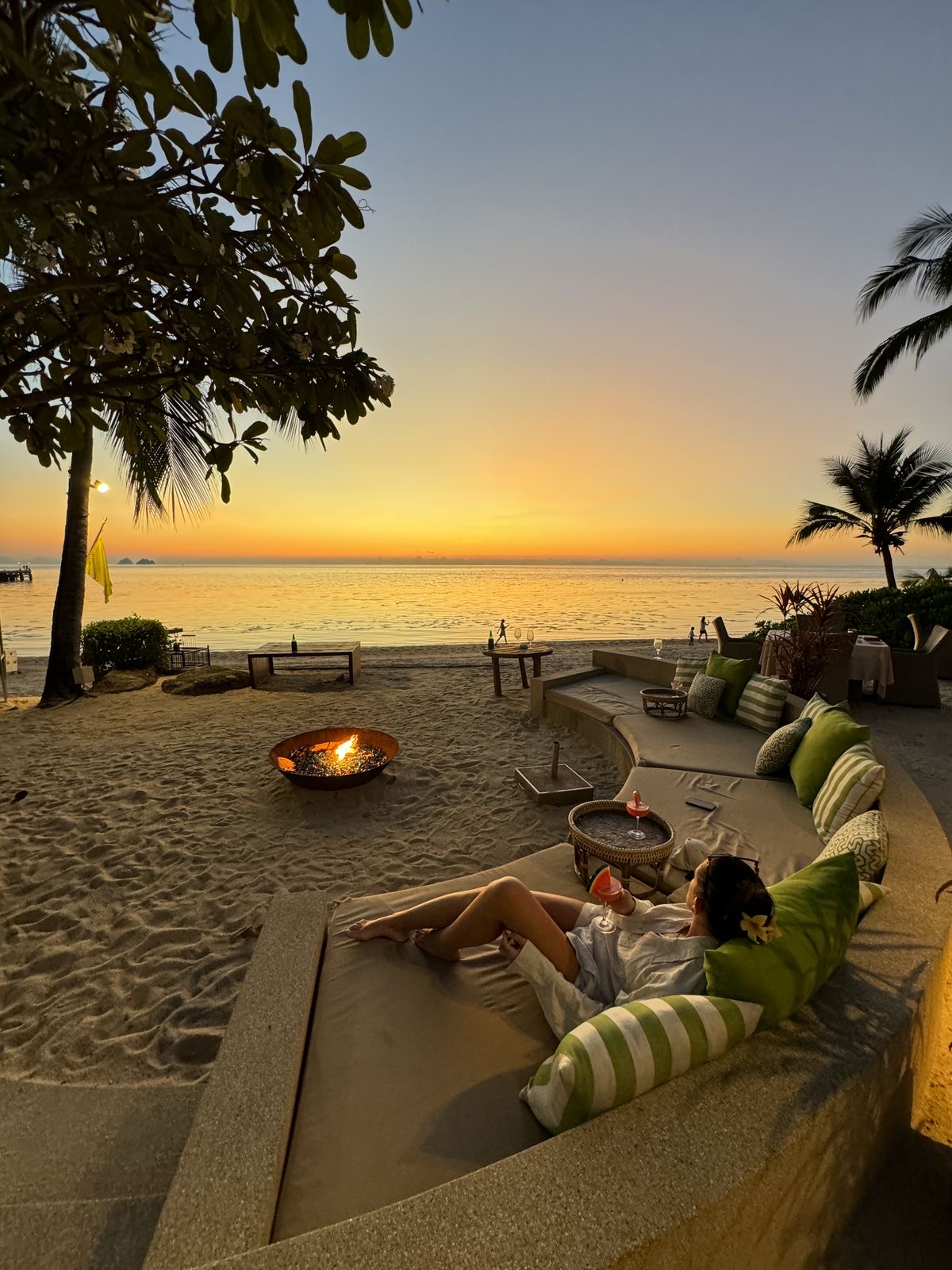 Beachfront lounge daybeds and a fire bowl on the sand facing a golden tropical sunset over the sea