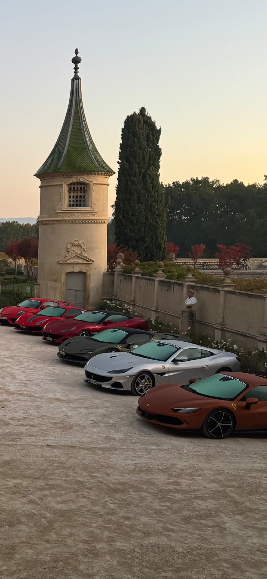A line of Ferraris parked at dusk in the courtyard of a southern French château beside a cypress tree