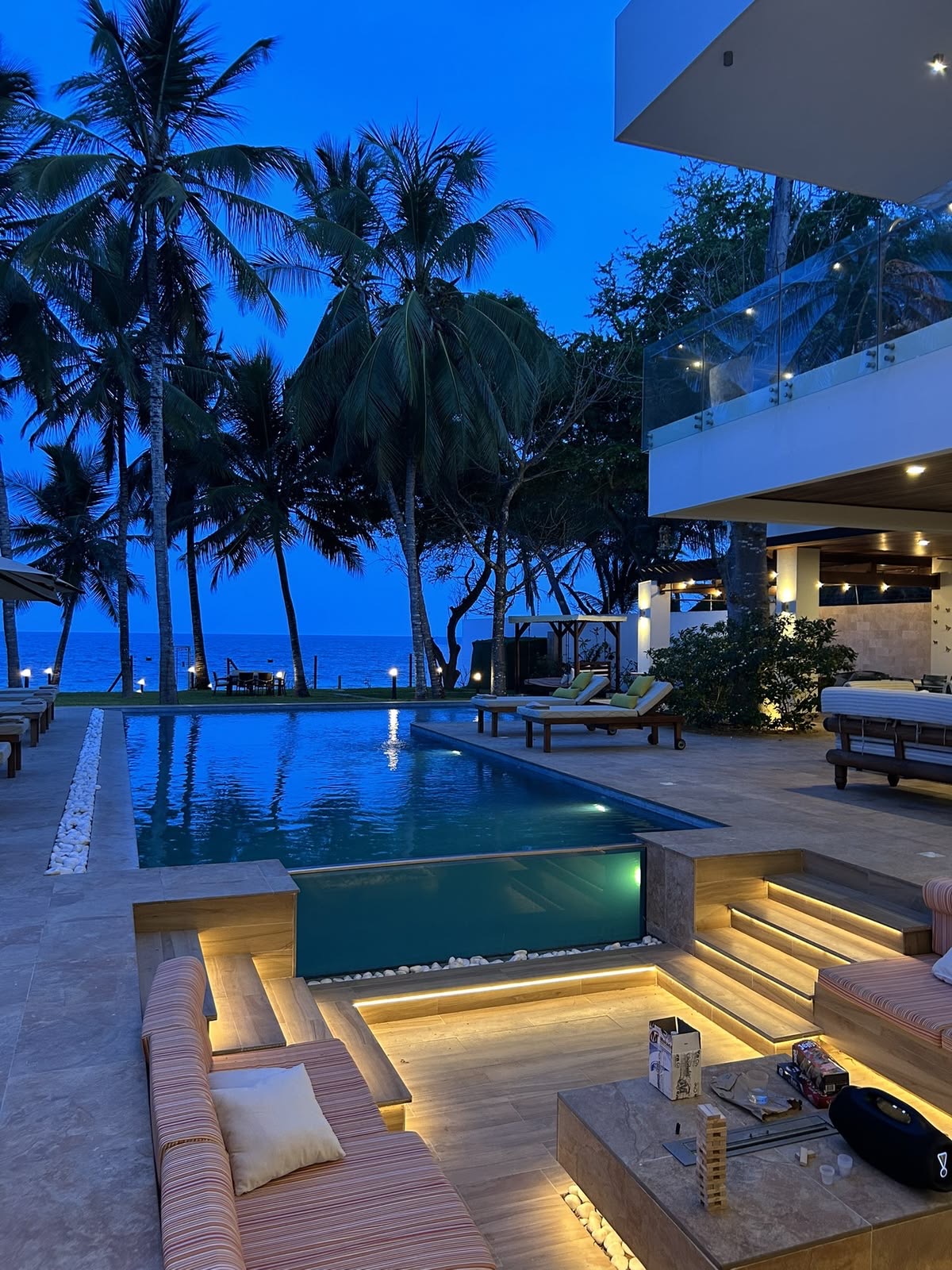 Tropical beachfront villa at dusk with a lit lap pool, palm trees and loungers facing the sea