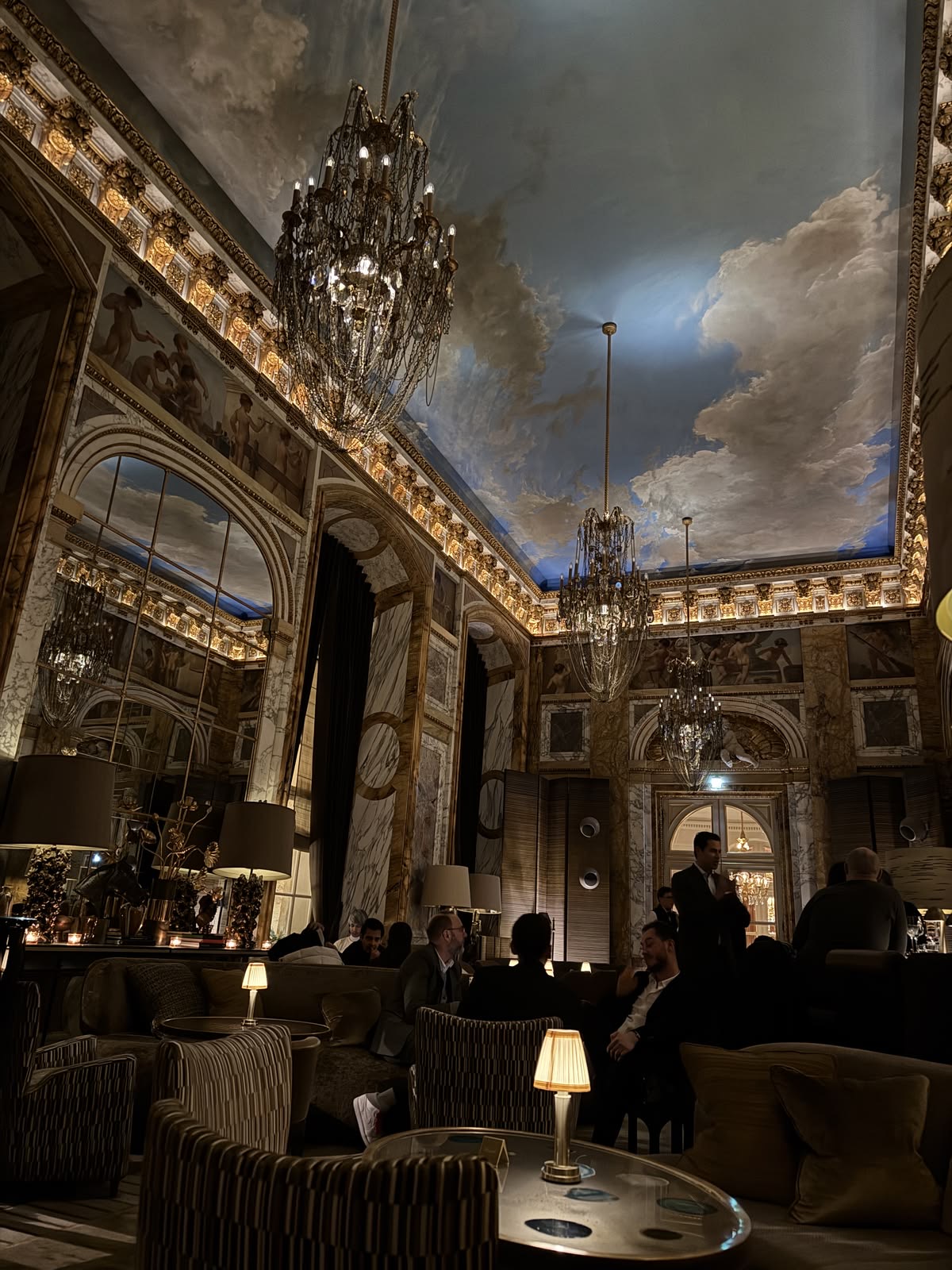 Grand Parisian palace salon with painted cloud ceiling, crystal chandeliers and lamplit tables
